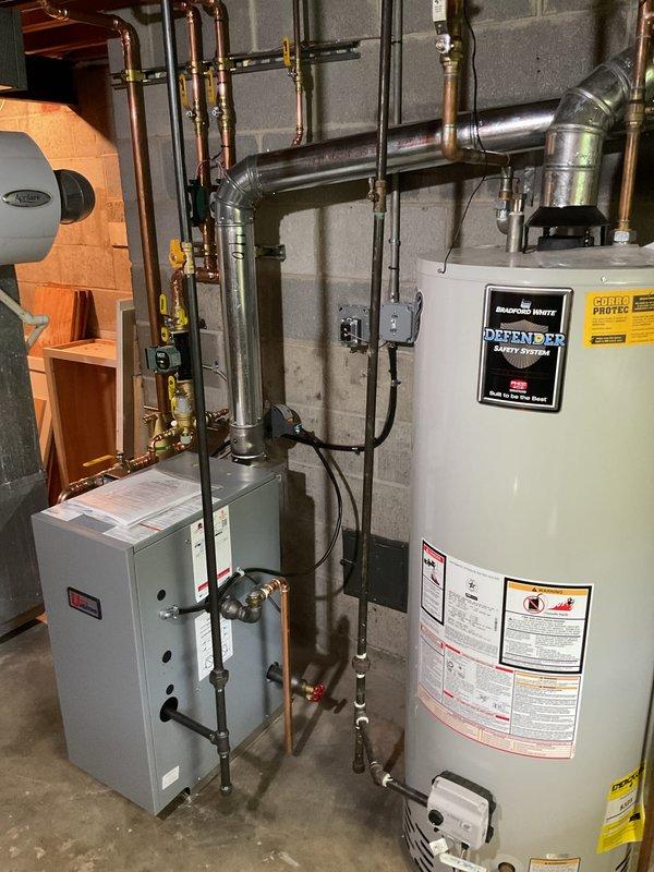 Completed full gas boiler installation in residential basement mechanical room, including all associated plumbing connections, copper piping network with brass fittings and shut-off valves, potable water expansion tank, and proper venting with metal ductwork. Installation includes Defender water heater integration with control systems, galvanized and copper pipe transitions, and complete HVAC infrastructure mounted to concrete block walls with proper support and code-compliant configuration. All equipment tested and operational, with service financing documentation completed and identification verified on file.