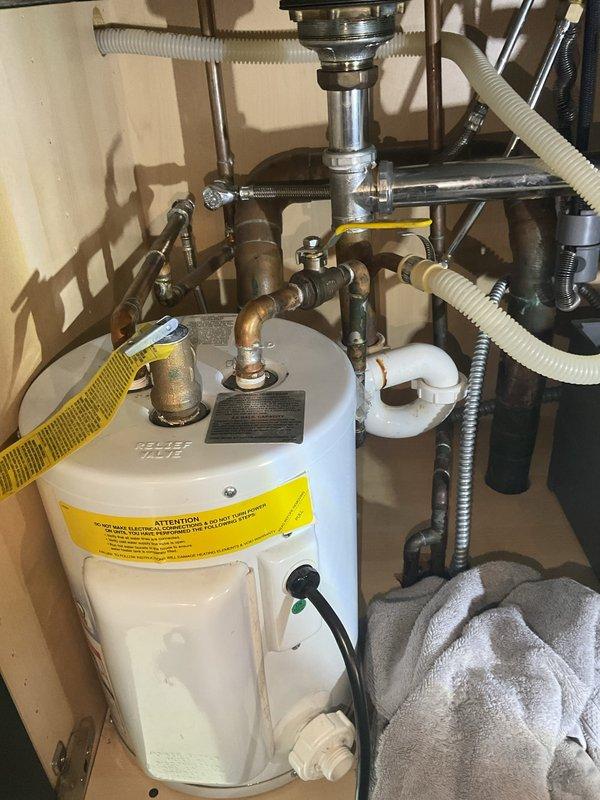 Conducted assessment for replacement of existing tank water heater with tankless system. Inspected current installation, which showed a conventional tank water heater with standard relief valve and copper supply lines in utility cabinet. Also evaluated kitchen sink plumbing requiring repair, noting oxidation on copper pipes and examining P-trap assembly and drain connections. Provided estimate for complete tankless conversion and separate pipe repair services.