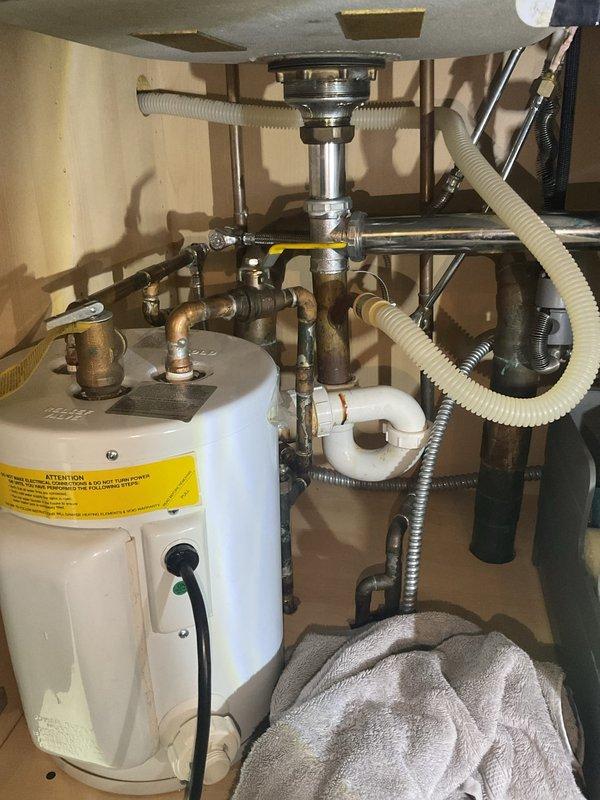Conducted assessment for replacement of existing tank water heater with tankless system. Inspected current installation, which showed a conventional tank water heater with standard relief valve and copper supply lines in utility cabinet. Also evaluated kitchen sink plumbing requiring repair, noting oxidation on copper pipes and examining P-trap assembly and drain connections. Provided estimate for complete tankless conversion and separate pipe repair services.