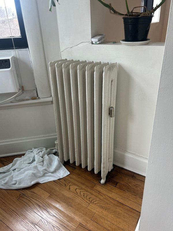Inspected cast iron radiator with reported valve leak near floor level. Found aged radiator valve showing signs of wear and discoloration. Valve is located at base of unit where it connects to supply line through wooden flooring with metal escutcheon plate.