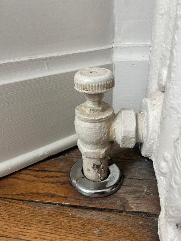 Inspected cast iron radiator with reported valve leak near floor level. Found aged radiator valve showing signs of wear and discoloration. Valve is located at base of unit where it connects to supply line through wooden flooring with metal escutcheon plate.