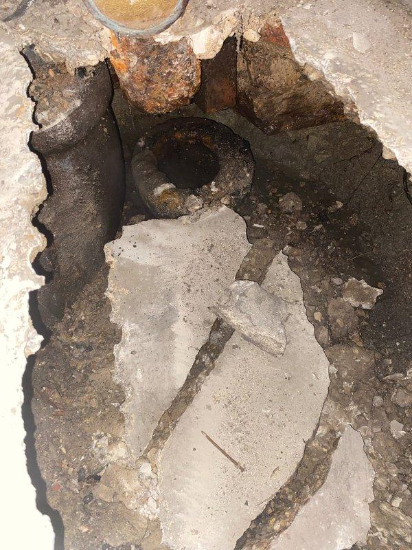Completed excavation of damaged concrete flooring to access severely corroded main drain line. Exposed circular metal pipe showed significant deterioration with extensive rust and surrounding soil contamination. Replaced deteriorated section with new main line drain pipe after clearing surrounding debris and compromised material.