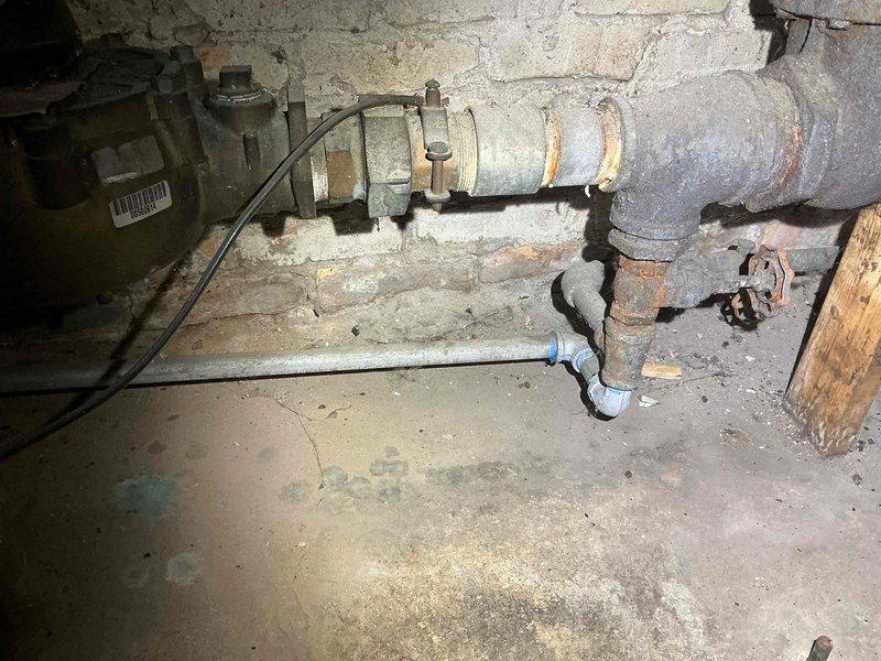 Conducted inspection of aging metal plumbing infrastructure in basement area, documenting significant oxidation and rust formation on pipe surfaces. Identified multiple connection points requiring maintenance, including T-junction and control valve. External inspection of sidewalk area completed with documentation of utility access point and building connection where vertical service pipe enters structure.
