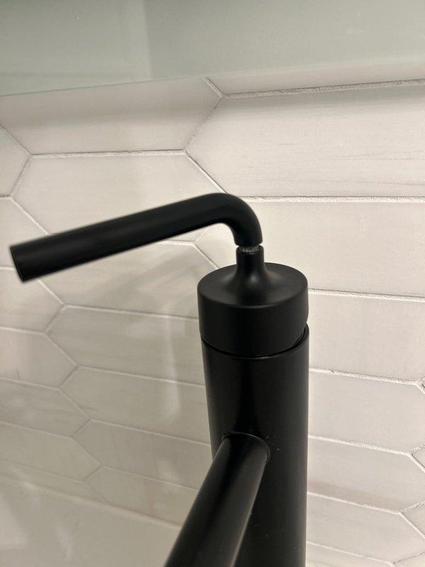 Completed installation of new matte black shower/tub fixtures as sold by Jacob. Mounted modern black faucet and dual shower system (handheld with flexible hose plus overhead rainfall component) against hexagonal white tile and marble-look surfaces. All fixtures properly installed with clean finish work evident in documentation photos.