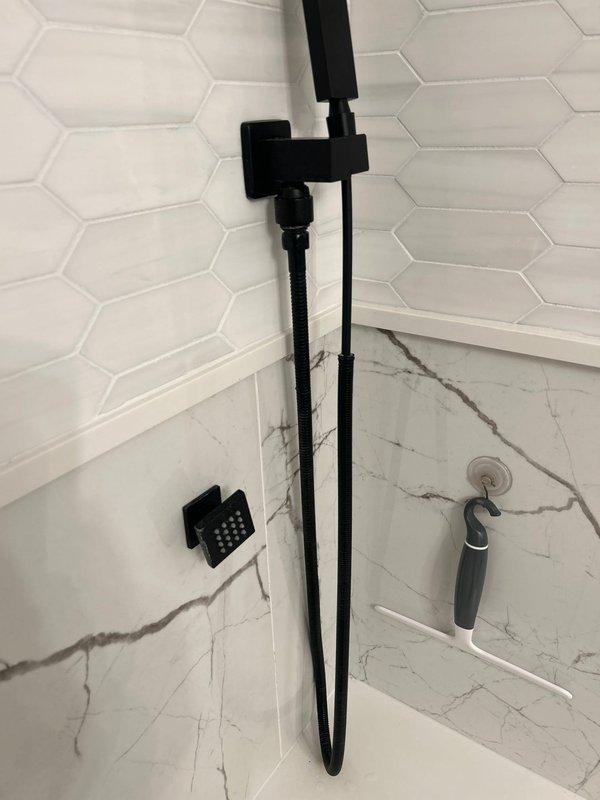 Completed installation of new matte black shower/tub fixtures as sold by Jacob. Mounted modern black faucet and dual shower system (handheld with flexible hose plus overhead rainfall component) against hexagonal white tile and marble-look surfaces. All fixtures properly installed with clean finish work evident in documentation photos.
