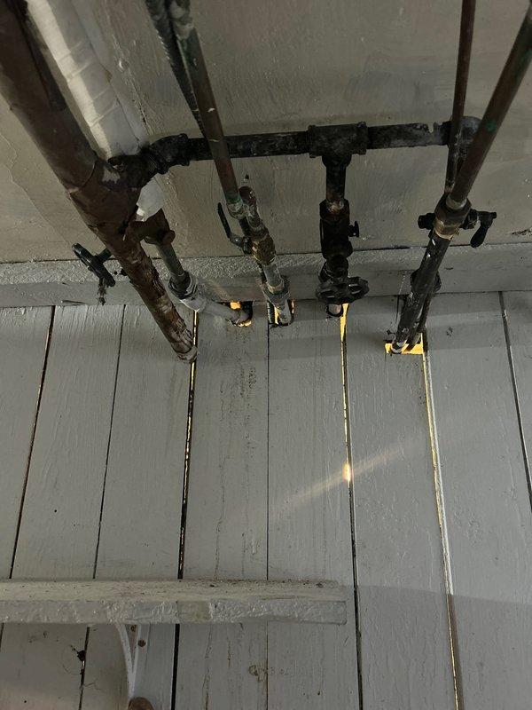 Located and identified water leak in basement originating from severely corroded pipe joints. Inspection revealed significant deterioration of connecting points between vertical and horizontal pipes with visible water damage and staining on surrounding ceiling/wall surfaces. Documented extensive corrosion and material degradation at critical junction points in the aging copper and metal pipe system.