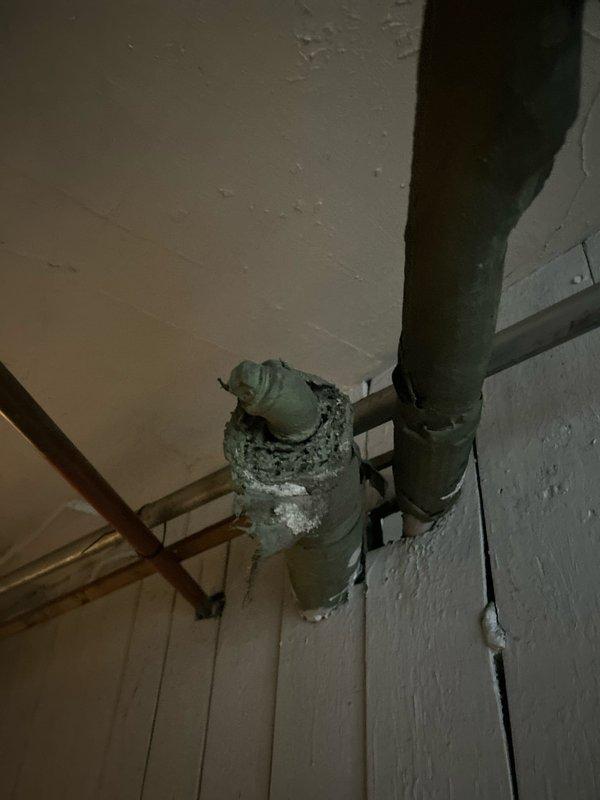 Located and identified water leak in basement originating from severely corroded pipe joints. Inspection revealed significant deterioration of connecting points between vertical and horizontal pipes with visible water damage and staining on surrounding ceiling/wall surfaces. Documented extensive corrosion and material degradation at critical junction points in the aging copper and metal pipe system.