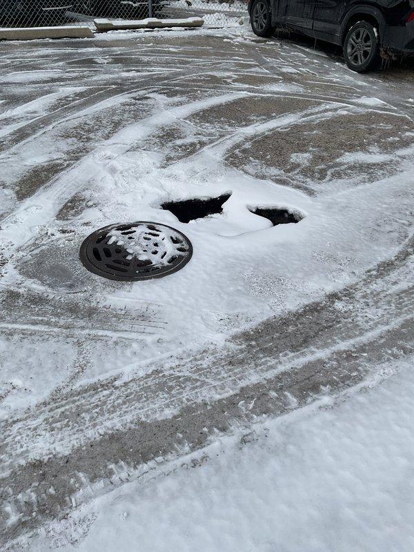 Conducted site inspection of reported sinkhole adjacent to manhole in snow-covered paved area. Visual assessment revealed potential undermining of pavement around drainage structure, with multiple depressions indicating possible pipe failure or soil erosion beneath surface. Recommended camera inspection to evaluate pipe integrity and determine extent of underground damage prior to excavation, repair/replacement of affected pipe sections, and backfilling to restore surface stability.