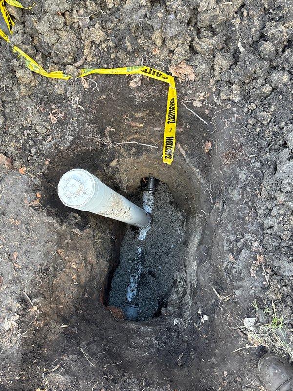 Excavated area to expose damaged underground plumbing line where a white PVC pipe connects to a black fitting. Job has been sold by Julio Basi and needs to be scheduled for completion of the repair to the compromised connection point showing signs of moisture/leakage.