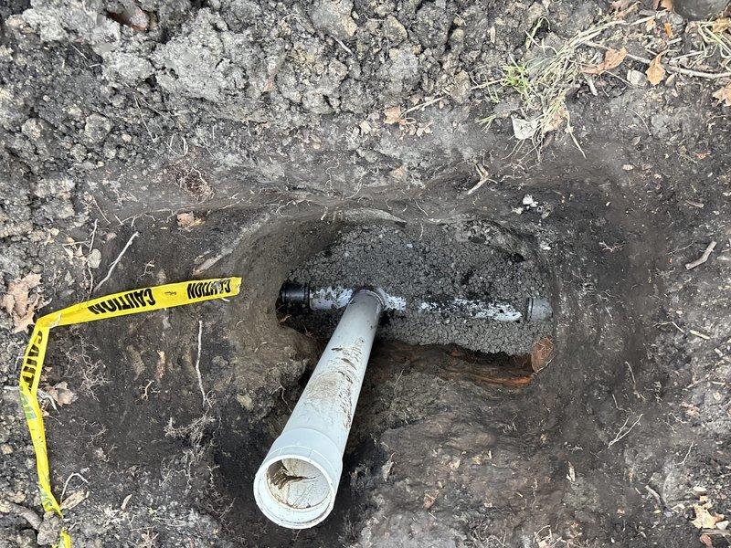 Excavated area to expose damaged underground plumbing line where a white PVC pipe connects to a black fitting. Job has been sold by Julio Basi and needs to be scheduled for completion of the repair to the compromised connection point showing signs of moisture/leakage.