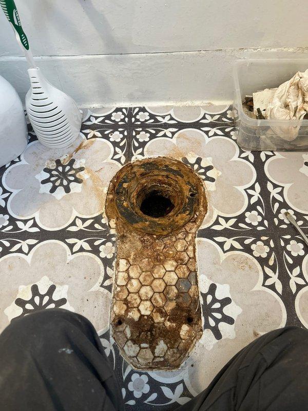 Removed severely corroded toilet flange and installed new two-piece white porcelain toilet in bathroom with decorative tile flooring. Secured unit properly with wall-mounted support bar for stability and confirmed proper functionality before completion.