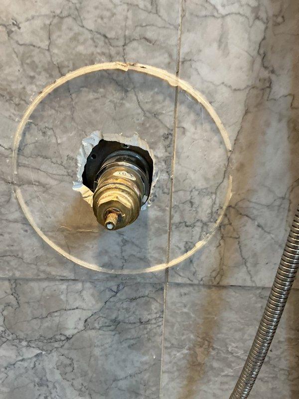 Accessed shower valve assembly through damaged trim ring in marble shower wall. Inspected brass valve components showing signs of aging. Documented condition of exposed plumbing fixture requiring replacement trim plate to properly finish installation.