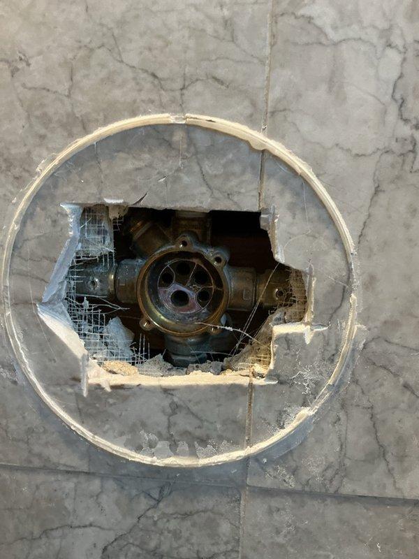 Accessed shower valve assembly through damaged trim ring in marble shower wall. Inspected brass valve components showing signs of aging. Documented condition of exposed plumbing fixture requiring replacement trim plate to properly finish installation.