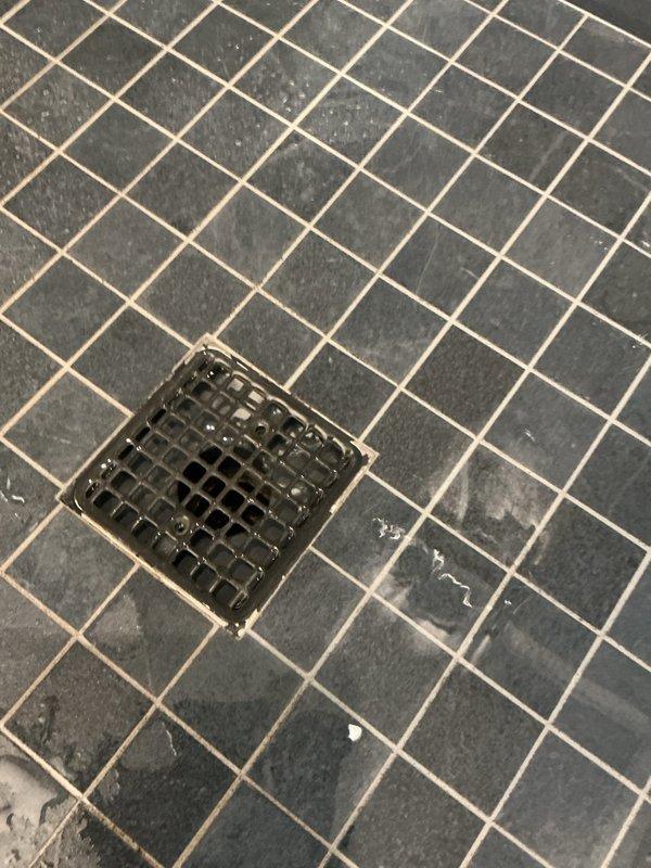 Responded to service call regarding shower drain clogging issue that began approximately 2 weeks prior to service date. Upon inspection, observed significant water marks and mineral deposits around the square metal drain cover in slate gray tiled shower floor. Cleared blockage and restored normal drainage function.