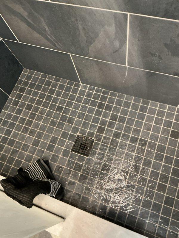 Responded to service call regarding shower drain clogging issue that began approximately 2 weeks prior to service date. Upon inspection, observed significant water marks and mineral deposits around the square metal drain cover in slate gray tiled shower floor. Cleared blockage and restored normal drainage function.