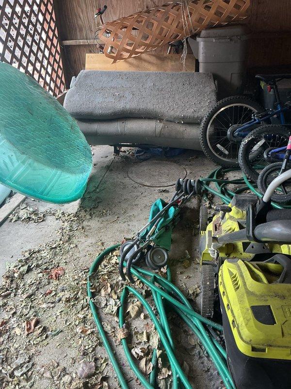 Inspected cluttered area under back porch to assess catch basin cleaning requirements. Found access points partially obstructed by stored bicycles, tools, and household items behind wooden lattice panels. Documented current conditions with photos for proper equipment planning and future service.