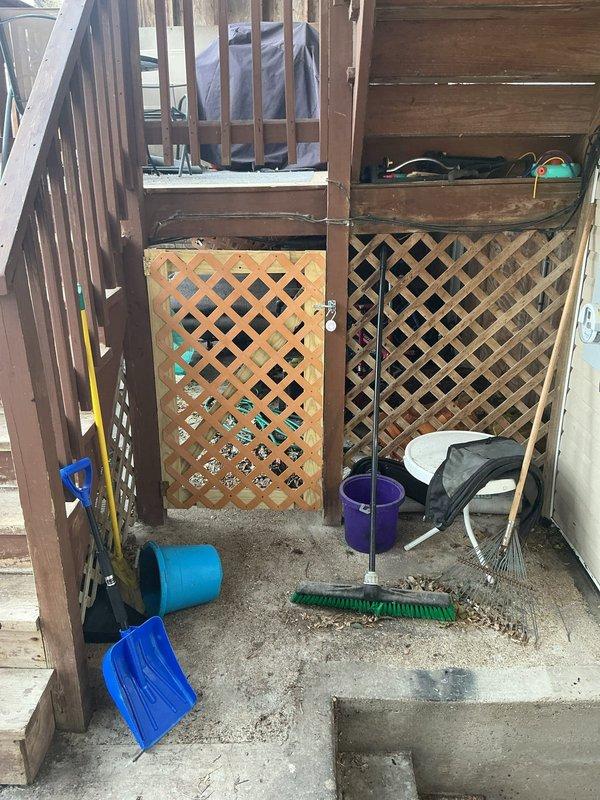 Inspected cluttered area under back porch to assess catch basin cleaning requirements. Found access points partially obstructed by stored bicycles, tools, and household items behind wooden lattice panels. Documented current conditions with photos for proper equipment planning and future service.