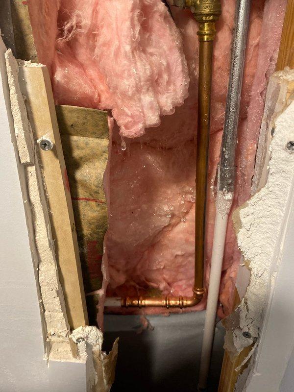 Responded to emergency call for burst water main supply line. Investigation revealed water intrusion at wall/floor junction with damaged insulation visible. Opened wall section to access affected area, exposing copper supply pipe with active leak at brass fitting. Repaired damaged section of pipe, replaced compromised fittings, and restored insulation before closing access point.