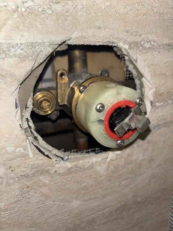 Accessed plumbing fixture through wall opening to perform repair on valve assembly as sold by Brian. Replaced deteriorated brass/copper fittings connected to the composite valve body with red collar. Installed new shower valve with gold-colored mounting plate, ensuring proper fitting within the circular wall opening. Photo documentation taken of both the access point and completed installation.