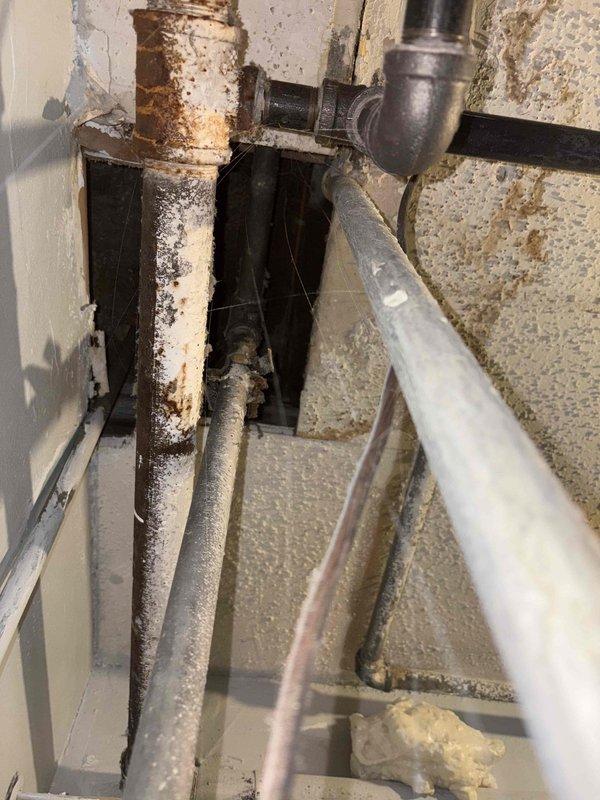 Inspected existing water heater system to provide replacement estimate. Observed significant corrosion and rust damage on vertical supply pipe, as well as deterioration in surrounding plumbing infrastructure. Identified potential water damage in utility area and documented current system configuration including pipe connections and flow indicators for accurate replacement specifications.