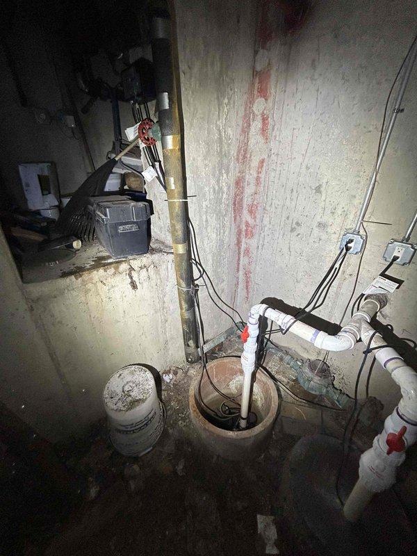 Responded to service call regarding sump pump backup battery alarm activation. Inspected the sump pump system in basement utility area, including battery backup unit, control panel with digital display, and associated PVC discharge piping. Determined battery required replacement and installed new backup battery to resolve persistent alarm issue.