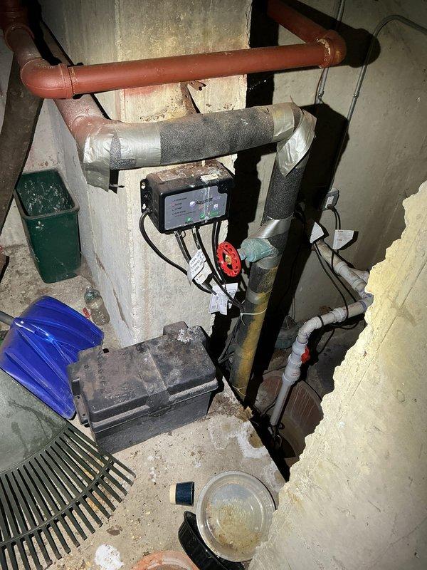 Responded to service call regarding sump pump backup battery alarm activation. Inspected the sump pump system in basement utility area, including battery backup unit, control panel with digital display, and associated PVC discharge piping. Determined battery required replacement and installed new backup battery to resolve persistent alarm issue.
