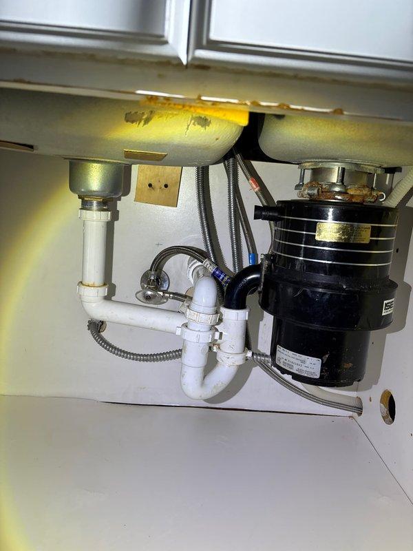 Completed wax ring replacement on basement toilet and inspected internal tank components. Examined kitchen faucet plumbing, noting P-trap configuration with PVC pipes and functional garbage disposal unit. Inspected copper pipe assembly with T-junction and verified proper connection of flexible metal conduit through wall.
