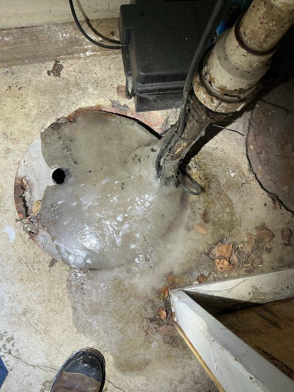 Inspected basement drainage system and identified a 5-6 year-old sump pump that has completely frozen and failed. Observed significant water pooling around the drain area with visible deterioration of the concrete floor and corrosion at pipe connection points. Documentation photos captured the damaged pump system and resulting water accumulation requiring complete replacement.