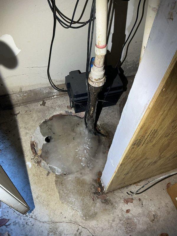 Inspected basement drainage system and identified a 5-6 year-old sump pump that has completely frozen and failed. Observed significant water pooling around the drain area with visible deterioration of the concrete floor and corrosion at pipe connection points. Documentation photos captured the damaged pump system and resulting water accumulation requiring complete replacement.