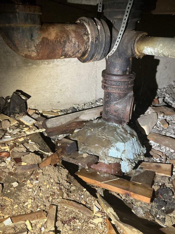 Inspected plumbing system in crawlspace and identified severely corroded cast iron pipe junction with extensive rust and deterioration. Documented multiple connection points showing significant oxidation damage, improper cement/mortar sealing at pipe bases, and accumulated debris around the plumbing infrastructure that requires attention.