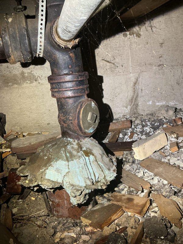 Inspected plumbing system in crawlspace and identified severely corroded cast iron pipe junction with extensive rust and deterioration. Documented multiple connection points showing significant oxidation damage, improper cement/mortar sealing at pipe bases, and accumulated debris around the plumbing infrastructure that requires attention.