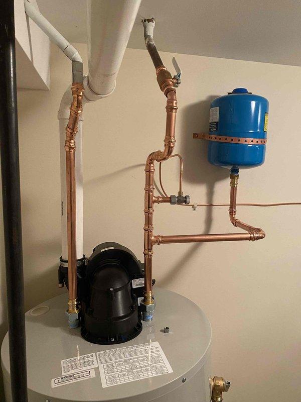 Completed installation of new water heater system with copper piping connections, including a pressure regulator and expansion tank mounted to the wall. System was properly fitted with necessary valves and connected to existing PVC discharge lines.