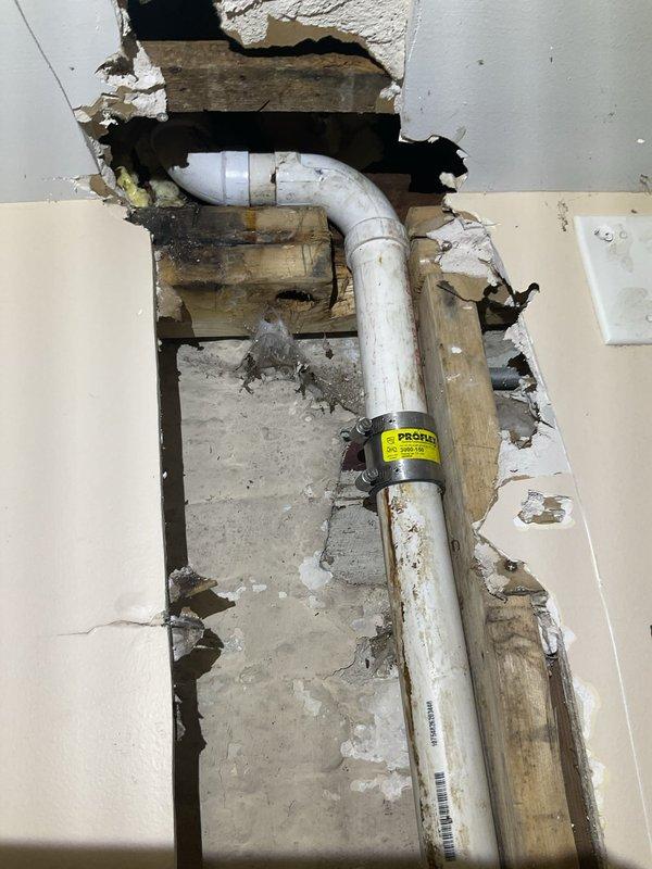 Located and identified a slow leak emanating from behind basement wall, requiring partial wall demolition to access the affected PVC piping. Inspected the exposed plumbing infrastructure, including elbow joint connections, and documented water damage to surrounding wall materials and wooden framing. Also examined kitchen area with dishwasher to verify no connection to the basement leak issue.
