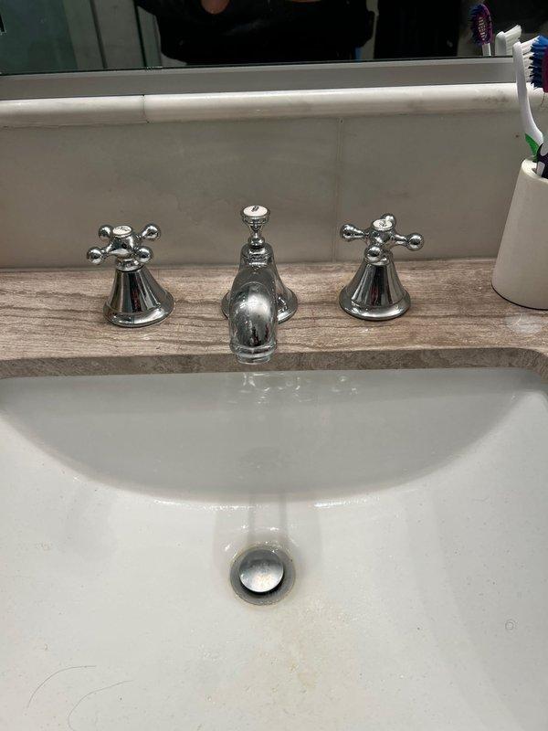 Replaced hot and cold cross-handle knobs on two bathroom sinks as requested by customer. Both sinks had three-piece faucet fixtures with chrome finish - one installed in a white porcelain sink with wood-patterned countertop, the other in a white square undermount basin with light brown marble countertop. Installation completed successfully with customer-provided matching replacement knobs.