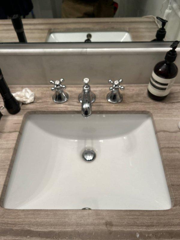 Replaced hot and cold cross-handle knobs on two bathroom sinks as requested by customer. Both sinks had three-piece faucet fixtures with chrome finish - one installed in a white porcelain sink with wood-patterned countertop, the other in a white square undermount basin with light brown marble countertop. Installation completed successfully with customer-provided matching replacement knobs.