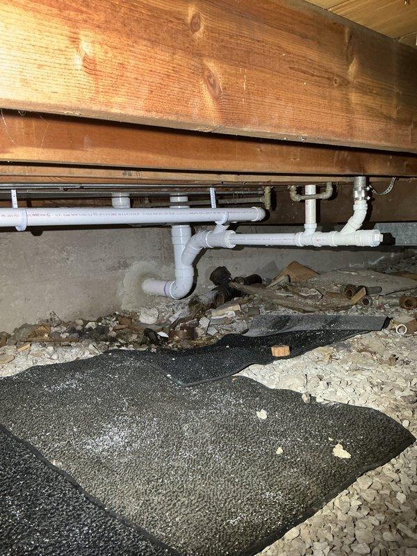 Conducted priority inspection of PVC drainage system in crawl space beneath wooden structure. Documented existing plumbing configuration including P-trap assembly and multiple pipe joints. Photographed exterior service area adjacent to foundation wall where plumbing exits structure through wooden enclosure for warranty documentation purposes.