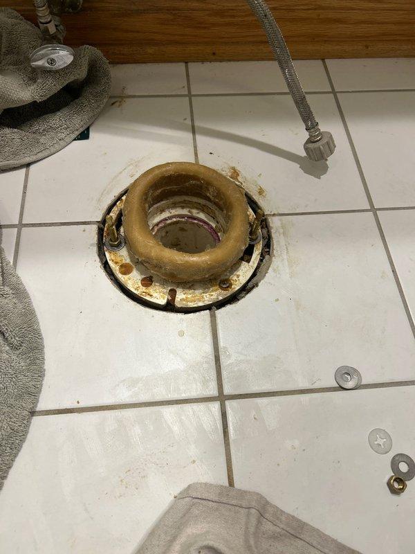 Technician removed toilet to inspect reported flush and clog issues, discovering a severely deteriorated toilet flange with extensive rust and mineral buildup. After assessment, replaced the corroded metal flange and installed new wax ring to restore proper seal and flushing functionality.