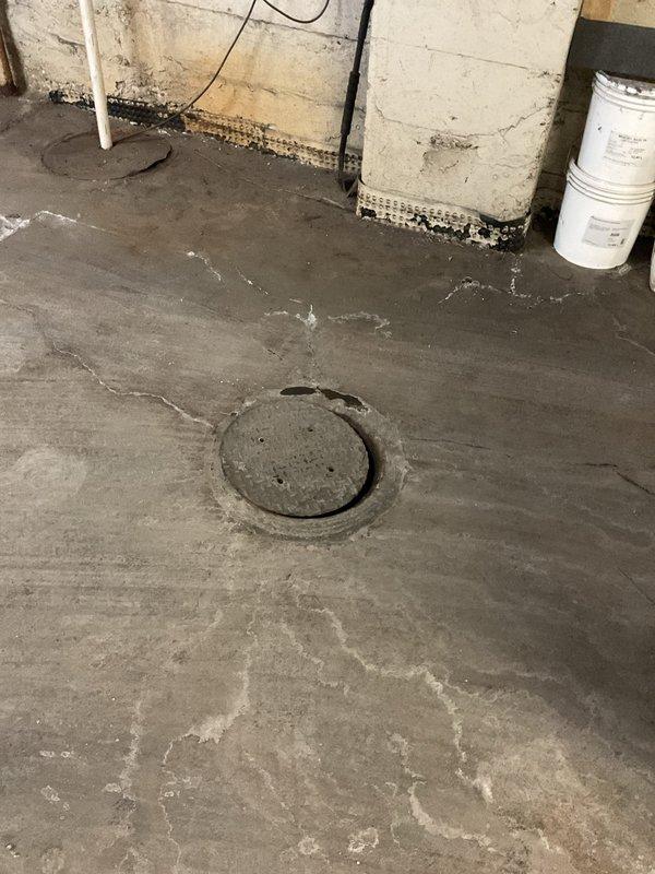 Conducted on-site assessment of damaged sewer cover in basement utility area. Measured existing circular access cover (approximately 17-18 inches in diameter) that showed significant deterioration and cracking. Documented surrounding concrete floor condition, which exhibited multiple settlement cracks and moisture issues at the wall junction. Provided customer with estimate for replacement cover as requested.