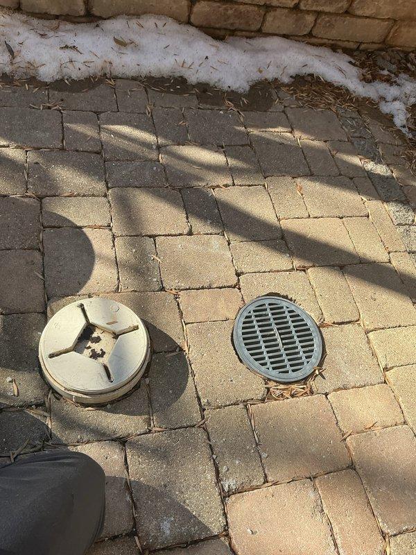 Performed inspection of residential catch basin system and drainage infrastructure. Identified two drain covers (one circular with cross-pattern design, one with standard grate pattern) installed in interlocking pavers, and examined water access utility cover. Provided assessment and recommendations for requested services including catch basin cleaning, mesh cap installation, and long-term options for drain tile modification, ejector pump installation, and potential catch basin removal.