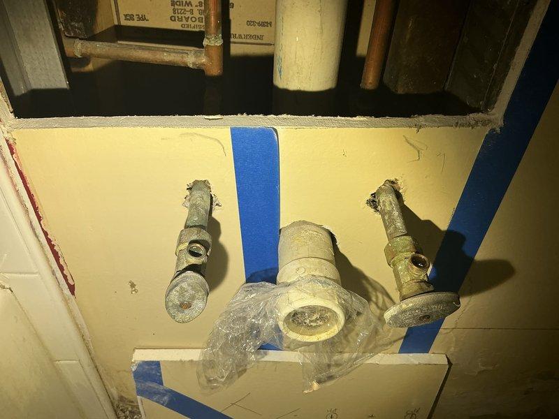 Conducted inspection of bathroom plumbing for requested drain line relocation and water line modifications during customer's remodel project. Observed existing fixtures showing significant corrosion and mineral buildup on valve stems, with damaged wall material surrounding connection points. Documented deteriorated PVC drain components and aging supply lines that will require replacement during the repositioning work.