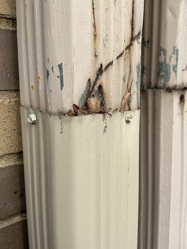 Inspected exterior downspout drainage system causing front yard flooding during rainfall. Identified significant deterioration at pipe seam with visible rust and corrosion. Found drain base partially clogged with accumulated debris including leaves, soil, and plant matter, preventing proper water flow from the downspout.