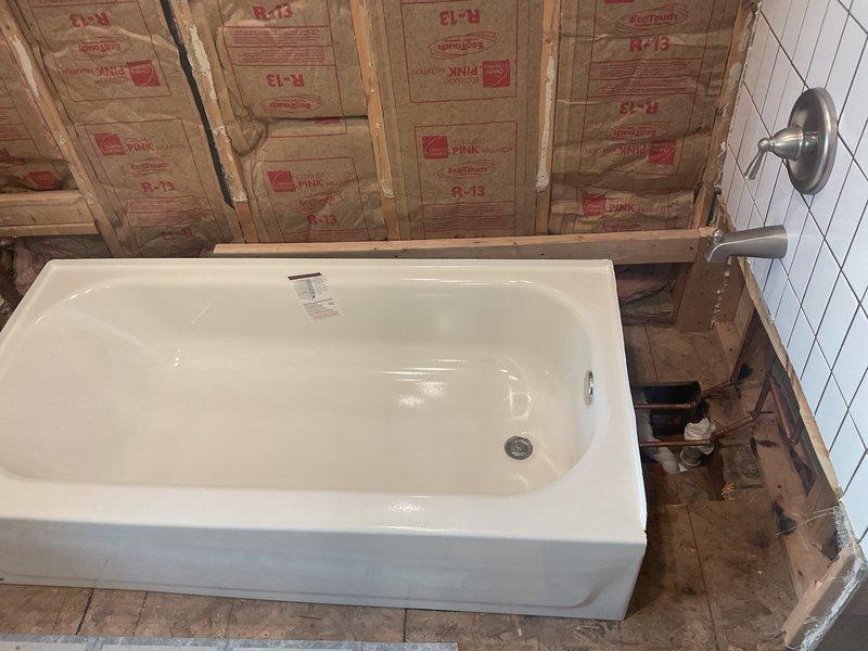 Successfully installed new porcelain bathtub with chrome drain on wooden subfloor and connected to existing plumbing system. Mounted stainless steel faucet fixture on adjacent tiled wall and ensured proper drain connection through floor cutout to PVC drain pipe below.