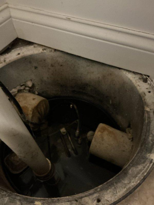 Inspected sump pump system with malfunctioning ejector pump float originally installed by Goode. Observed aged sump pump that has never been replaced, with visible signs of deterioration including dirt accumulation and discoloration. Examined PVC drainage configuration with vertical pipe secured to wall via metal mounting bracket, noting purple primer at connection points.