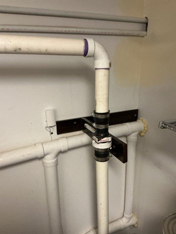 Inspected sump pump system with malfunctioning ejector pump float originally installed by Goode. Observed aged sump pump that has never been replaced, with visible signs of deterioration including dirt accumulation and discoloration. Examined PVC drainage configuration with vertical pipe secured to wall via metal mounting bracket, noting purple primer at connection points.