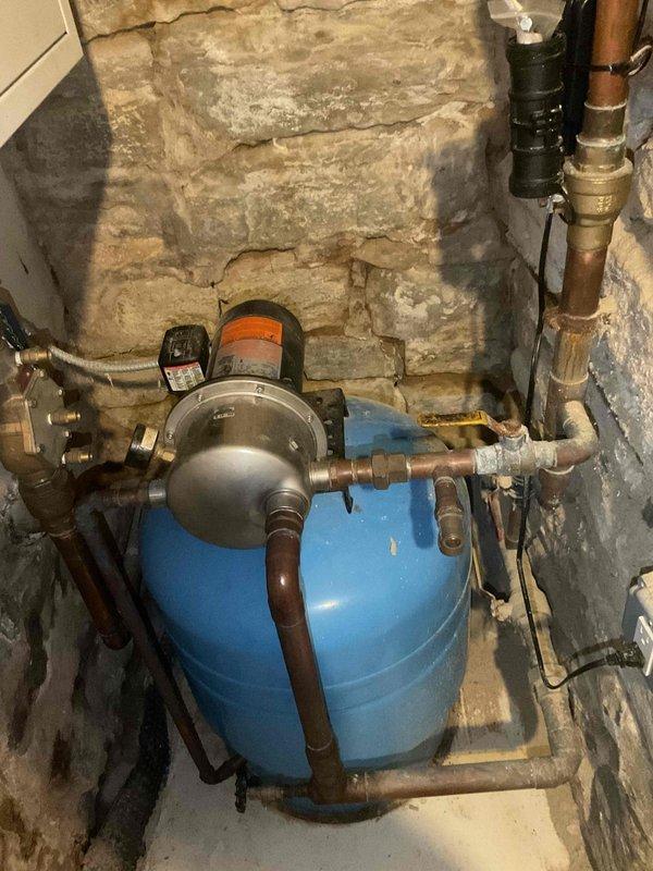 Conducted on-site inspection of existing well water system, documenting current configuration including aged pressure tank, pump assembly, and associated copper piping. Photographed complete installation showing the blue pressure tank with connected valves, fittings, and the pump/motor unit mounted against stone foundation wall. Prepared estimate for recommended piping upgrades to address visible oxidation and aging components.