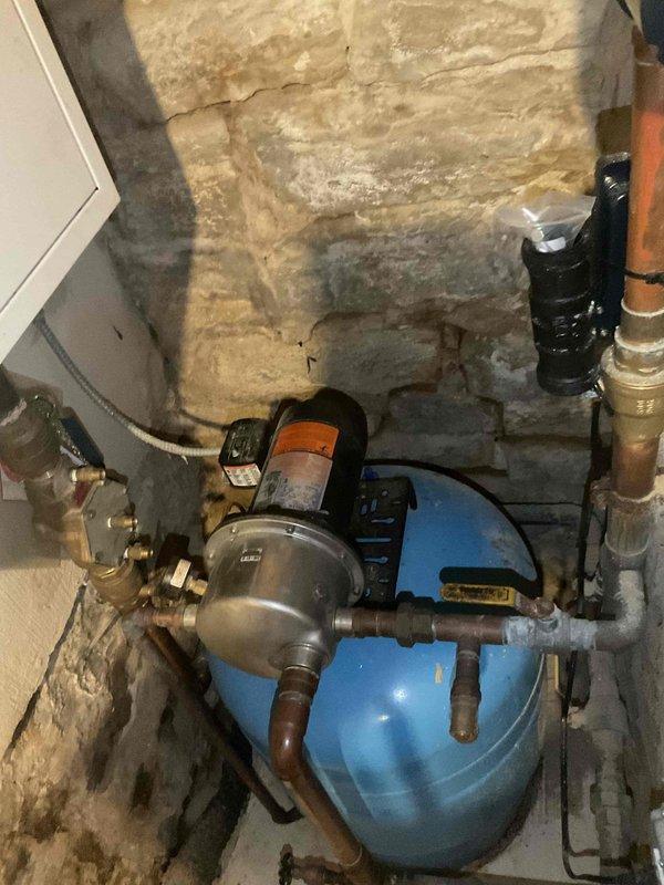 Conducted on-site inspection of existing well water system, documenting current configuration including aged pressure tank, pump assembly, and associated copper piping. Photographed complete installation showing the blue pressure tank with connected valves, fittings, and the pump/motor unit mounted against stone foundation wall. Prepared estimate for recommended piping upgrades to address visible oxidation and aging components.