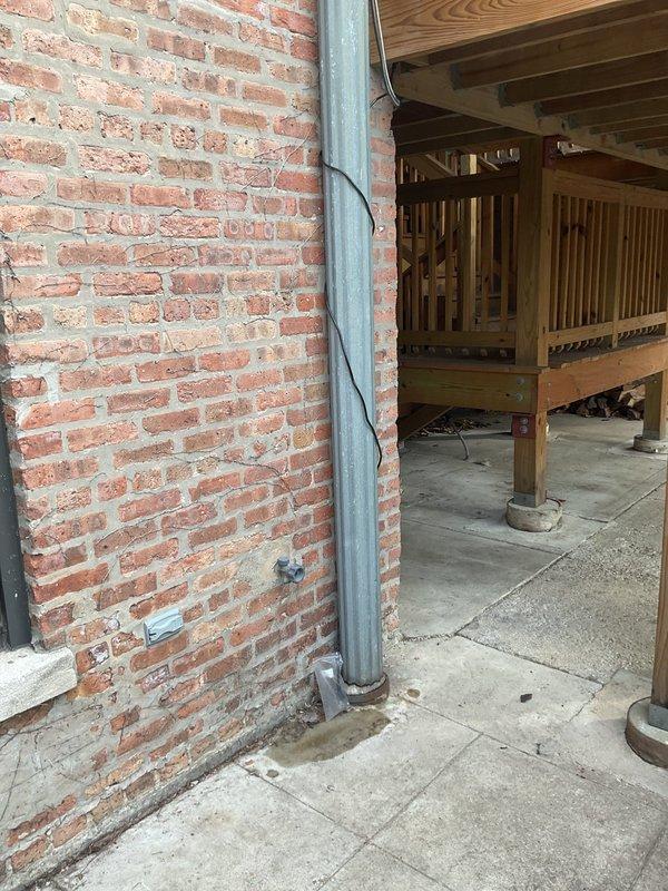 Inspected exterior downspout that was reported spraying at seams and leaking. Visual examination confirmed issue at connection points along brick wall where galvanized metal downspout is secured with brackets. Documented condition with photographs showing the affected area adjacent to wooden deck structure and patio area. Repaired compromised seams to prevent further water leakage and ensure proper drainage function.