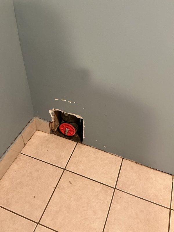 Located and inspected water leak in basement bathroom wall, identified source at a corner junction where plumbing valve is located. Created access opening in drywall to expose the red valve fixture and evaluate the extent of damage; documented condition with photographs for repair assessment.