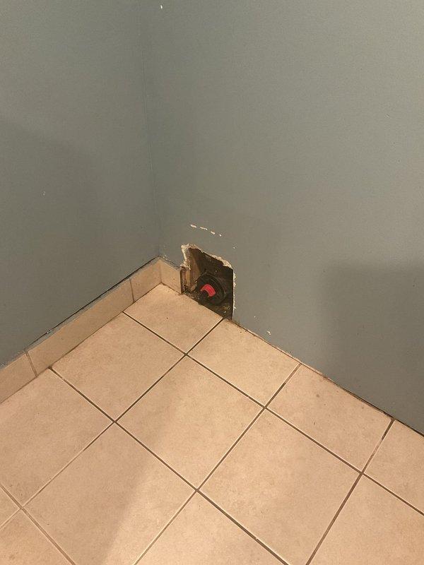 Located and inspected water leak in basement bathroom wall, identified source at a corner junction where plumbing valve is located. Created access opening in drywall to expose the red valve fixture and evaluate the extent of damage; documented condition with photographs for repair assessment.