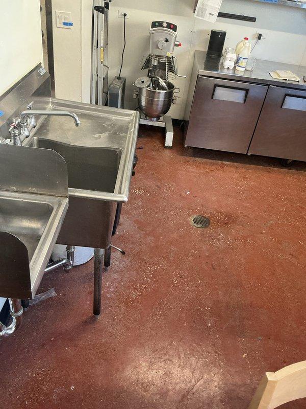 Responded to customer request for hydrojetting estimate after experiencing water backups from floor drains when operating certain sinks. Inspected commercial kitchen area including stainless steel sink units, visible floor drains, and adjacent plumbing infrastructure. Identified grease trap requiring maintenance and documented evidence of water damage and oxidation in the utility area where pipe assemblies connect to drainage system.